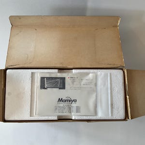 May include: An open rectangular cardboard box containing white styrofoam packaging inserts and a white paper document from Mamiya Camera Co. Ltd. The document features black text and a small technical diagram printed on its surface.