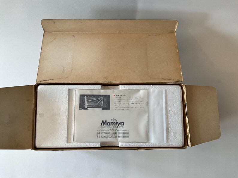 May include: An open rectangular cardboard box containing white styrofoam packaging inserts and a white paper document from Mamiya Camera Co. Ltd. The document features black text and a small technical diagram printed on its surface.