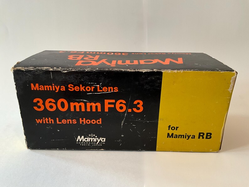 May include: A rectangular black and yellow cardboard box for a Mamiya Sekor Lens 360mm F6.3 with Lens Hood for Mamiya RB. The box features orange text reading Mamiya Sekor Lens 360mm F6.3 with Lens Hood and the Mamiya Camera Co. Ltd. logo.