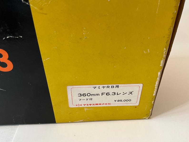 May include: A yellow and black box featuring a white rectangular label with Japanese text that reads Mamiya RB, 360mm F6.3 lens, and includes a hood. The label also displays the manufacturer name Mamiya Optical Co., Ltd. in Japanese characters.