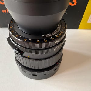 May include: A black camera lens featuring a wide, flared lens hood at the top. The barrel displays white and orange numerical markings for focus and aperture settings, with textured grip rings for manual adjustments and a small red alignment dot.