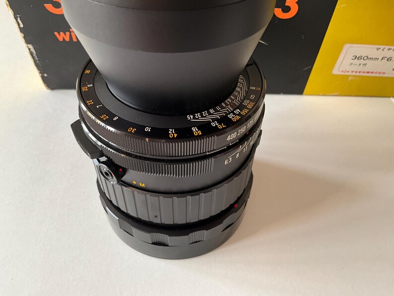 May include: A black camera lens featuring a wide, flared lens hood at the top. The barrel displays white and orange numerical markings for focus and aperture settings, with textured grip rings for manual adjustments and a small red alignment dot.