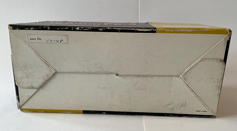 May include: A rectangular cardboard box with a black, white, and yellow color scheme. The top panel features the text "Lens No. 10148" in a white box. The bottom right corner includes the text "Made in Japan" and handwritten markings.