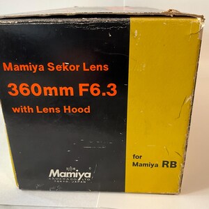 May include: A rectangular cardboard product box with a black and yellow color scheme. The text reads Mamiya Sekor Lens 360mm F6.3 with Lens Hood for Mamiya RB and Mamiya Camera Co. Ltd. Tokyo, Japan. The box shows signs of wear on the edges.