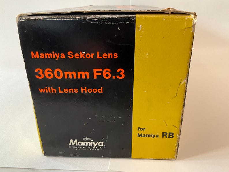May include: A rectangular cardboard product box with a black and yellow color scheme. The text reads Mamiya Sekor Lens 360mm F6.3 with Lens Hood for Mamiya RB and Mamiya Camera Co. Ltd. Tokyo, Japan. The box shows signs of wear on the edges.