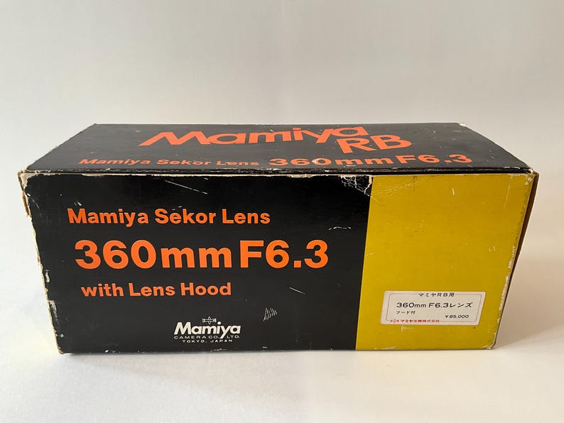May include: A black and yellow rectangular cardboard box for a Mamiya RB Mamiya Sekor Lens 360mm F6.3 with Lens Hood. The box features orange text reading Mamiya RB, Mamiya Sekor Lens, 360mm F6.3, and with Lens Hood, with a white label on the side.