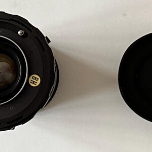 May include: A black camera lens with a circular glass element, metal mounting ring, and small red and green alignment dots. A small yellow sticker with the letters BH is attached to the side. A separate black circular lens cap is placed nearby.