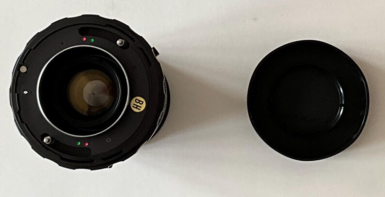 May include: A black camera lens with a circular glass element, metal mounting ring, and small red and green alignment dots. A small yellow sticker with the letters BH is attached to the side. A separate black circular lens cap is placed nearby.