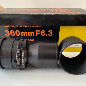 May include: A black Mamiya RB 360mm F6.3 camera lens with a separate black lens hood, positioned in front of its original black and yellow cardboard box. The box features the text Mamiya RB, Mamiya Sekor Lens, and 360mm F6.3 in orange.