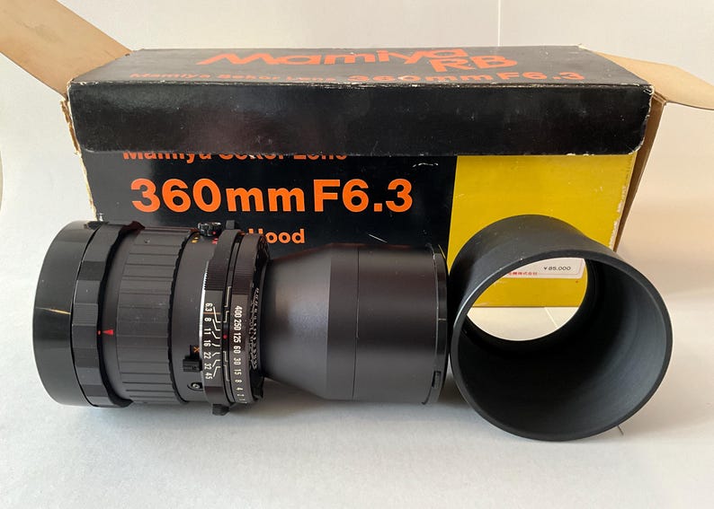 May include: A black Mamiya RB 360mm F6.3 camera lens with a separate black lens hood, positioned in front of its original black and yellow cardboard box. The box features the text Mamiya RB, Mamiya Sekor Lens, and 360mm F6.3 in orange.
