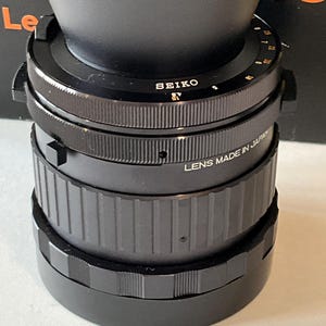 May include: A black Seiko camera lens featuring a textured focus ring and a metal barrel with white engraved text that reads SEIKO and LENS MADE IN JAPAN. The lens includes a rotating aperture ring with numerical markings and a knurled grip surface.