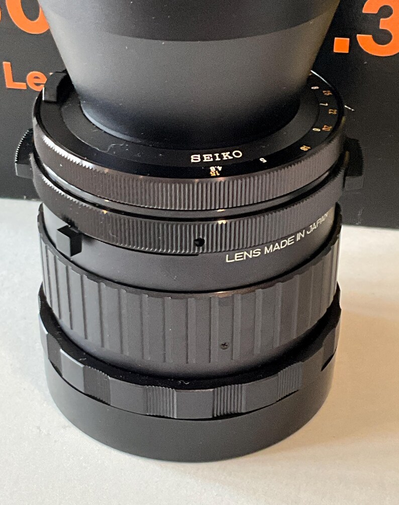 May include: A black Seiko camera lens featuring a textured focus ring and a metal barrel with white engraved text that reads SEIKO and LENS MADE IN JAPAN. The lens includes a rotating aperture ring with numerical markings and a knurled grip surface.