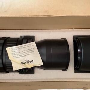 May include: A black Mamiya camera lens resting in a white styrofoam insert within a cardboard box. A small, off-white instruction paper with black text is attached to the lens, featuring the Mamiya logo and instructions for lens mounting.