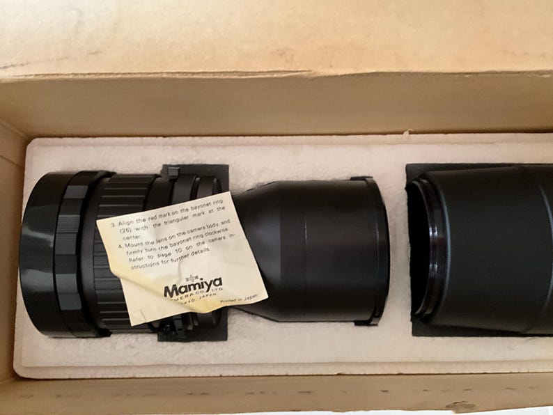 May include: A black Mamiya camera lens resting in a white styrofoam insert within a cardboard box. A small, off-white instruction paper with black text is attached to the lens, featuring the Mamiya logo and instructions for lens mounting.