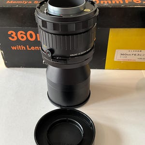 May include: A black Mamiya RB 360mm F6.3 camera lens with a matching lens hood and original black and orange box. The lens features a textured focus ring, a silver mount, and the text "LENS MADE IN JAPAN" printed on the side of the barrel.