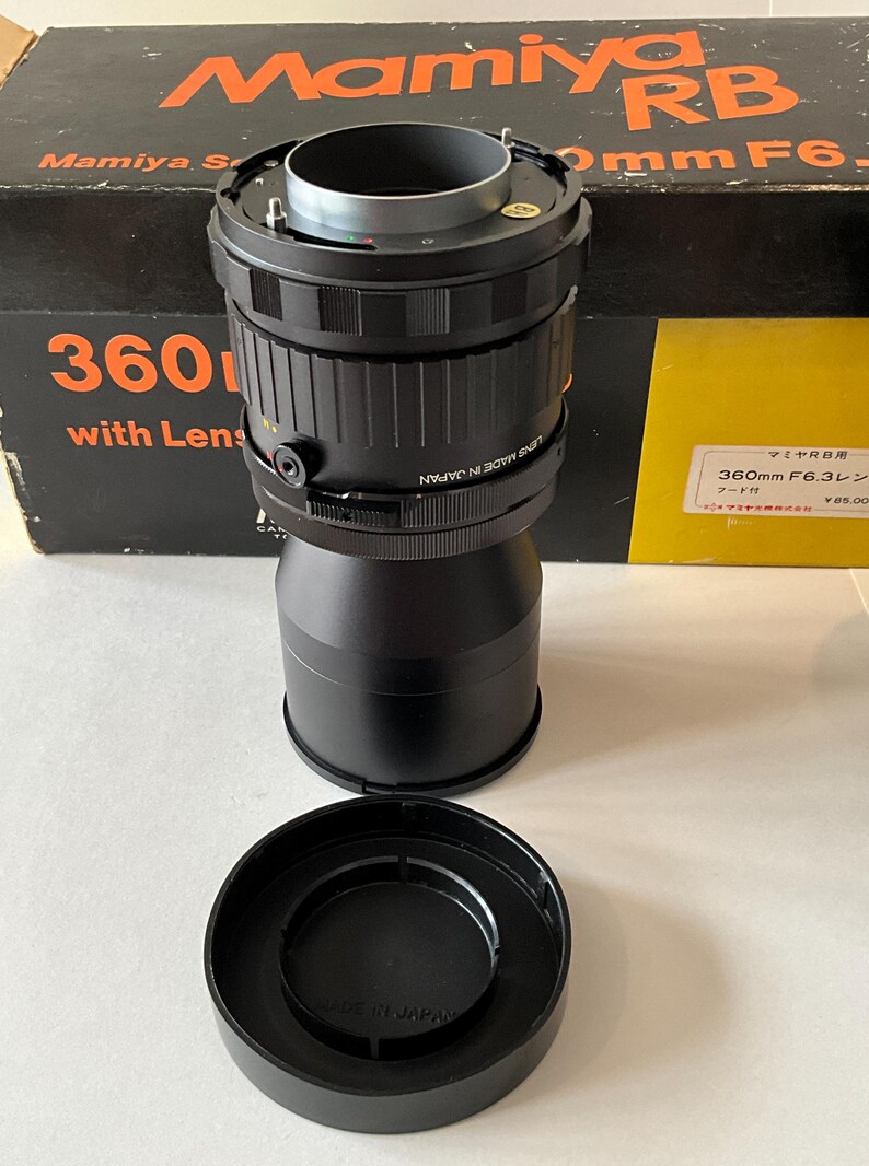 May include: A black Mamiya RB 360mm F6.3 camera lens with a matching lens hood and original black and orange box. The lens features a textured focus ring, a silver mount, and the text "LENS MADE IN JAPAN" printed on the side of the barrel.