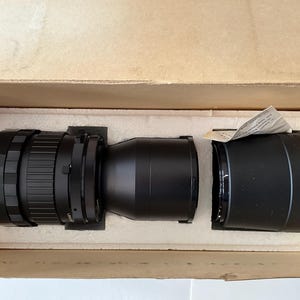 May include: A black camera lens with a ribbed focus ring and a separate black lens hood, both resting inside a tan cardboard box with white foam inserts. A small piece of white paper with printed text is tucked into the lens hood.