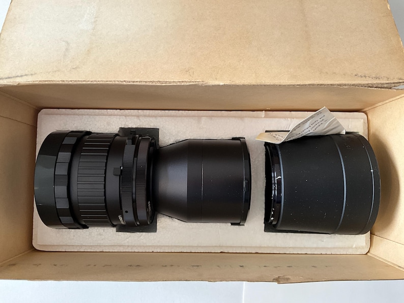 May include: A black camera lens with a ribbed focus ring and a separate black lens hood, both resting inside a tan cardboard box with white foam inserts. A small piece of white paper with printed text is tucked into the lens hood.