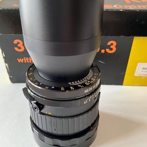 May include: A black camera lens with a wide barrel and multiple adjustment rings featuring white numerical markings for focus and aperture settings. The lens is positioned in front of a black and yellow box with orange text that reads 360mm 6.3.