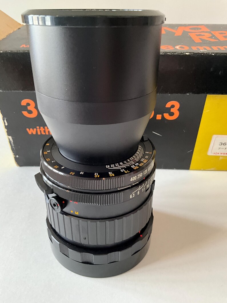 May include: A black camera lens with a wide barrel and multiple adjustment rings featuring white numerical markings for focus and aperture settings. The lens is positioned in front of a black and yellow box with orange text that reads 360mm 6.3.