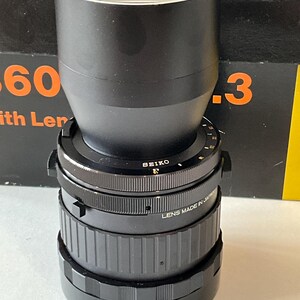 May include: A black Mamiya camera lens featuring a Seiko shutter mechanism. The lens barrel is marked with the text No. 10148 and LENS MADE IN JAPAN. It has a cylindrical shape with textured focus and aperture rings, and a wide, smooth front lens hood.