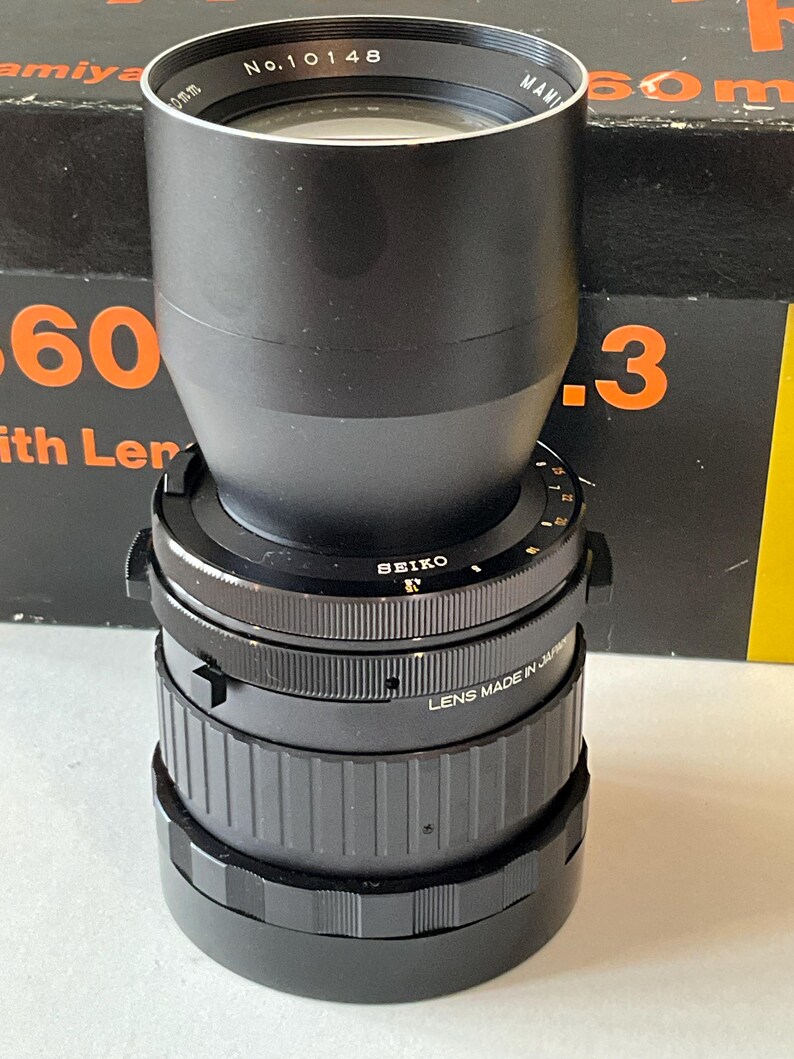 May include: A black Mamiya camera lens featuring a Seiko shutter mechanism. The lens barrel is marked with the text No. 10148 and LENS MADE IN JAPAN. It has a cylindrical shape with textured focus and aperture rings, and a wide, smooth front lens hood.