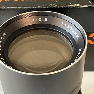 May include: A black Mamiya Sekor camera lens with a silver rim. The front element is surrounded by text reading Mamiya Sekor 1:6.3 f=360mm. The lens is positioned in front of a black box with orange text, partially visible in the background.