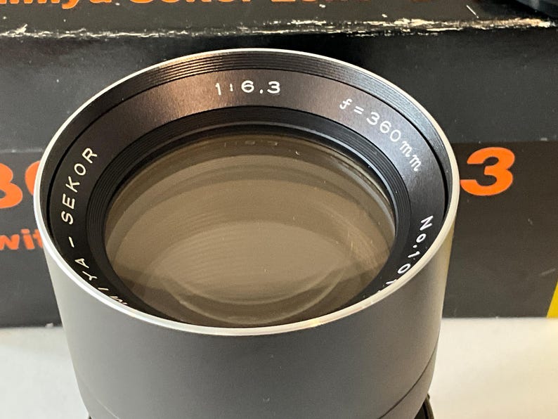 May include: A black Mamiya Sekor camera lens with a silver rim. The front element is surrounded by text reading Mamiya Sekor 1:6.3 f=360mm. The lens is positioned in front of a black box with orange text, partially visible in the background.