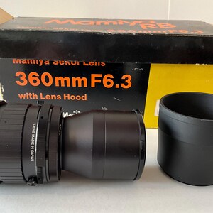 May include: A black Mamiya Sekor 360mm F6.3 camera lens with a separate black lens hood, positioned in front of its original cardboard box. The box is black and yellow with orange text reading Mamiya RB, Mamiya Sekor Lens, and 360mm F6.3.
