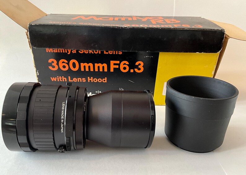 May include: A black Mamiya Sekor 360mm F6.3 camera lens with a separate black lens hood, positioned in front of its original cardboard box. The box is black and yellow with orange text reading Mamiya RB, Mamiya Sekor Lens, and 360mm F6.3.