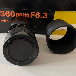 May include: A black camera lens with a front cap and a separate black lens hood, positioned in front of a product box featuring the text 360mm F6.3 with L. The lens has a cylindrical body with textured grip rings and a smooth, circular front element.