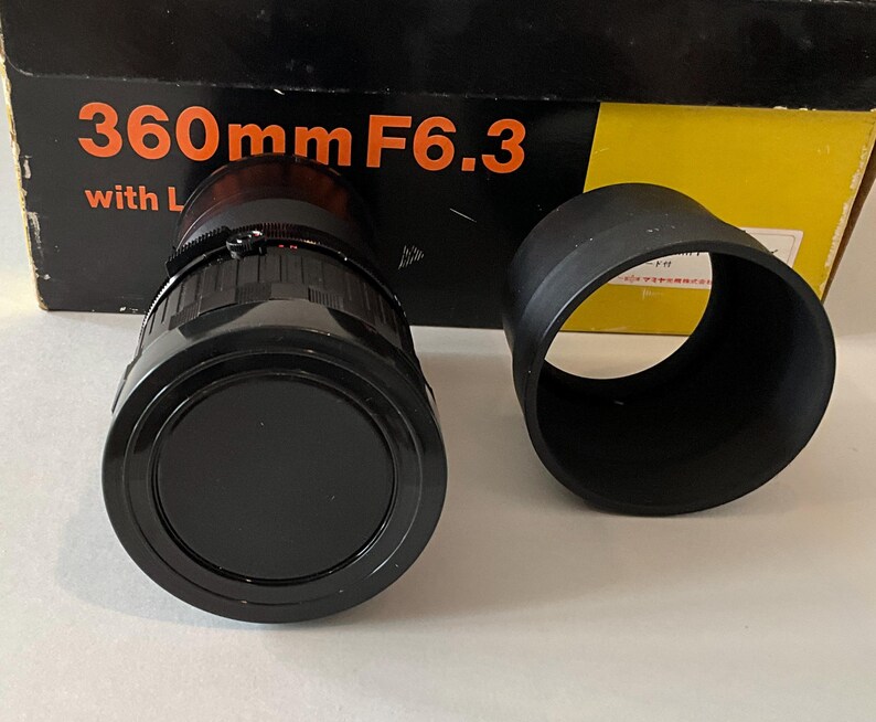 May include: A black camera lens with a front cap and a separate black lens hood, positioned in front of a product box featuring the text 360mm F6.3 with L. The lens has a cylindrical body with textured grip rings and a smooth, circular front element.