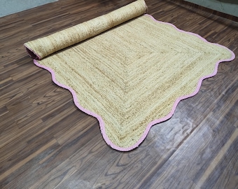 May include: A rolled-up, beige jute rug with a pink scalloped border. The rug is partially unrolled, revealing its textured weave and the pink trim. The rug is on a wooden floor.