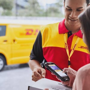 以下が含まれることがあります： 赤、黄、黒の制服を着たDHLの配達員が、顧客に荷物を渡しています。顧客はスマートフォンの画面を見て配達を確認しています。背景には黄色のDHLのバンがあります。