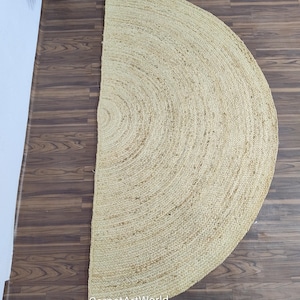 May include: A half-circle jute rug in a natural beige color. The rug has a woven texture with concentric circular patterns. The rug is on a dark brown wooden floor. The text "CarpetArtWorld" is visible.