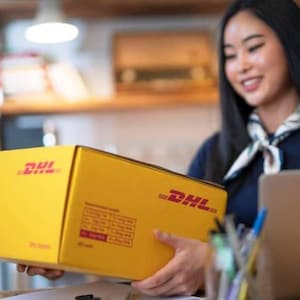 以下が含まれることがあります： 赤色のDHLロゴが入った黄色のDHL配送ボックス。箱は人が持っています。箱には情報が記載されたラベルが付いています。背景はぼやけています。
