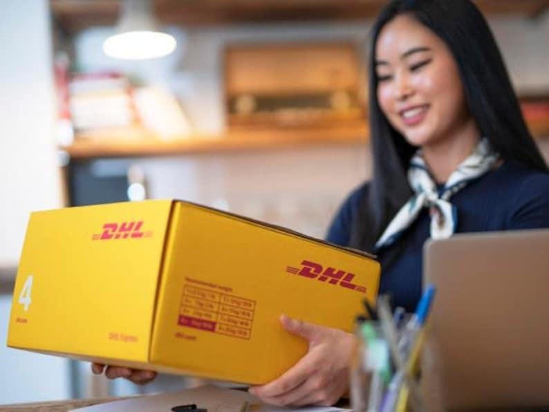 以下が含まれることがあります： 赤色のDHLロゴが入った黄色のDHL配送ボックス。箱は人が持っています。箱には情報が記載されたラベルが付いています。背景はぼやけています。