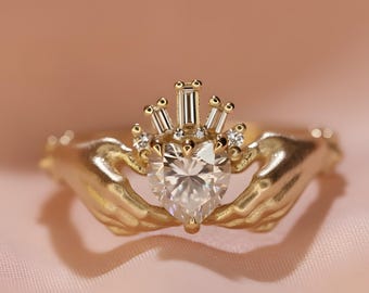 May include: A gold-colored Claddagh ring with a heart-shaped gemstone held by two hands. The gemstone is surrounded by smaller stones and a crown design. The ring is set against a pink background.