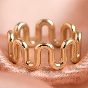 May include: A gold-toned ring featuring a continuous, repeating wavy or serpentine pattern that forms the band. The metal surface is smooth and reflective, creating a consistent, rhythmic design that encircles the entire circumference of the ring.