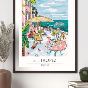 Op de afbeelding: Een ingelijste kunstafdruk met een caf&eacute;sc&egrave;ne aan zee in Saint-Tropez, Frankrijk. De illustratie toont een gele parasol, tafels met drankjes en een uitzicht op de zee met een boot. De afdruk bevat de tekst "ST. TROPEZ FRANCE".