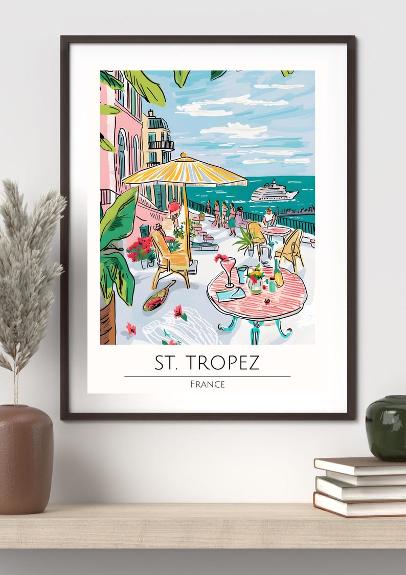 Op de afbeelding: Een ingelijste kunstafdruk met een caf&eacute;sc&egrave;ne aan zee in Saint-Tropez, Frankrijk. De illustratie toont een gele parasol, tafels met drankjes en een uitzicht op de zee met een boot. De afdruk bevat de tekst "ST. TROPEZ FRANCE".