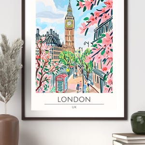 Op de afbeelding: Een ingelijste kunstprint van Londen, VK, met de Big Ben en een rode telefooncel. De illustratie heeft een kleurrijke, grillige stijl met roze bloesems en een blauwe lucht. Het woord "LONDON" staat onder de afbeelding.