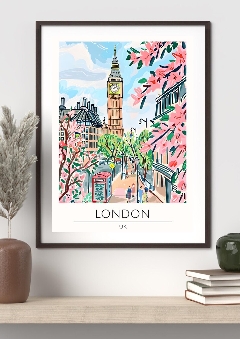 Op de afbeelding: Een ingelijste kunstprint van Londen, VK, met de Big Ben en een rode telefooncel. De illustratie heeft een kleurrijke, grillige stijl met roze bloesems en een blauwe lucht. Het woord "LONDON" staat onder de afbeelding.