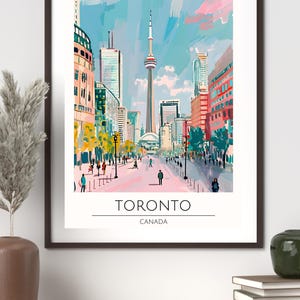 Op de afbeelding: Een ingelijste kunstdruk van Toronto, Canada, met een levendige straatsc&egrave;ne met de CN Tower. Het kunstwerk gebruikt een kleurrijke, impressionistische stijl met een blauwe en roze lucht, en de tekst "TORONTO CANADA" onderaan.