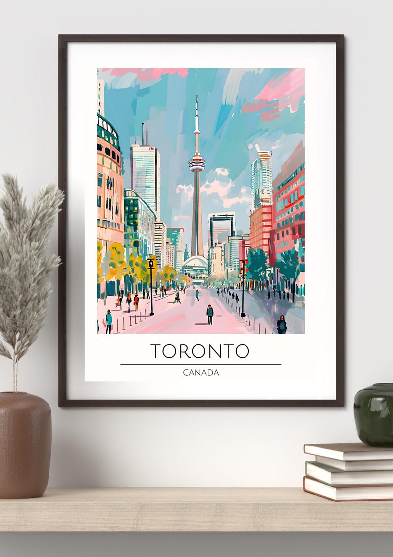 Op de afbeelding: Een ingelijste kunstdruk van Toronto, Canada, met een levendige straatsc&egrave;ne met de CN Tower. Het kunstwerk gebruikt een kleurrijke, impressionistische stijl met een blauwe en roze lucht, en de tekst "TORONTO CANADA" onderaan.