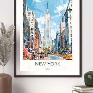 Op de afbeelding: Een ingelijste kunstafdruk van New York City, met het Empire State Building. Het kunstwerk heeft een aquarel-stijl, met een blauwe lucht en gele taxi's. De tekst "NEW YORK USA" staat onderaan.