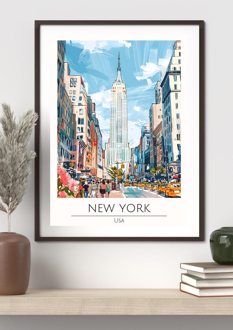 Op de afbeelding: Een ingelijste kunstafdruk van New York City, met het Empire State Building. Het kunstwerk heeft een aquarel-stijl, met een blauwe lucht en gele taxi's. De tekst "NEW YORK USA" staat onderaan.