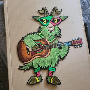 May include: An embroidered patch featuring a green goat wearing 3D-style glasses with the number 3 on each lens. The goat is playing a brown acoustic guitar and wearing pink and green leg warmers, a green patterned collar, and a green strap.