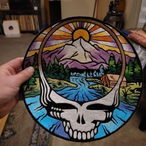 May include: A circular embroidered patch featuring a stylized skull outline framing a landscape scene. The design includes a yellow sun, purple mountains, green pine trees, a blue river, and a small tan tent, all rendered in vibrant thread colors.