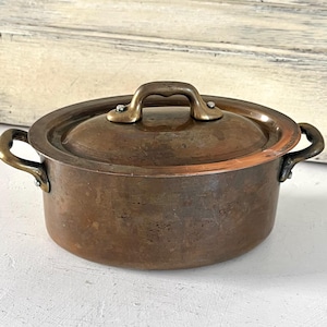 Puede incluir: Una olla redonda de cobre con tapa a juego, que cuenta con dos asas laterales y un asa central en la tapa. La superficie metálica muestra una pátina oscura y desgastada con signos visibles de envejecimiento y oxidación en el cuerpo.